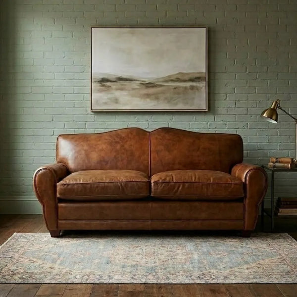 A brown leather loveseat sofa placed in a styled room with a green brick wall, a framed landscape painting above, a patterned rug underneath, and a floor lamp to the right.