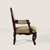 An ornate vintage-style armchair with a carved dark brown wooden frame and light beige upholstered seat and backrest, shown from a side profile against a solid light beige background.