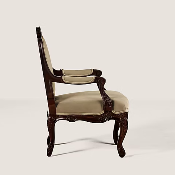 An ornate vintage-style armchair with a carved dark brown wooden frame and light beige upholstered seat and backrest, shown from a side profile against a solid light beige background.