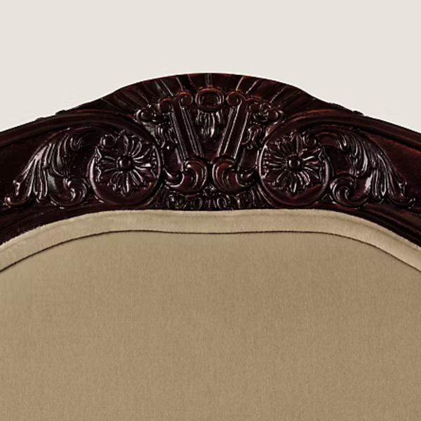 Close-up of an upholstered chair back showing a dark carved wooden headboard with ornate scrollwork and a beige fabric seat.
