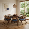 A 8-piece dining set featuring a round dark wood table and six matching armchairs with woven cane backs and blue cushions, styled in a bright dining room with a woven pendant light and large window view.