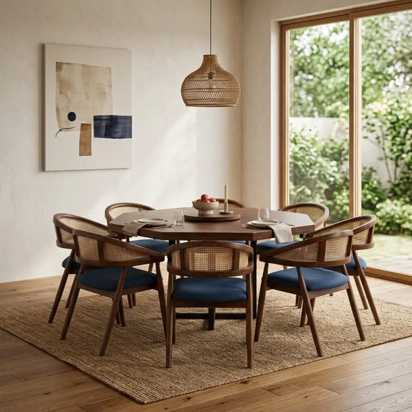 A 8-piece dining set featuring a round dark wood table and six matching armchairs with woven cane backs and blue cushions, styled in a bright dining room with a woven pendant light and large window view.