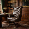 A classic brown leather tufted office chair with a high back and swivel base, placed in a traditional wood-paneled study next to a desk and lamp.