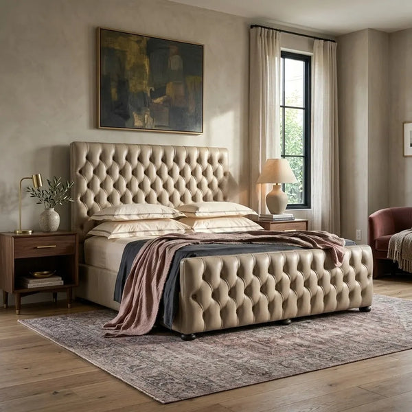 A beige tufted upholstered bed frame with a matching headboard and footboard, styled in a warm, neutral-toned bedroom with wood floors, nightstands, and a patterned rug.