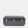 A black leather or faux-leather trunk with gold-tone metal hardware, corner protectors, and rivets, photographed against a plain light gray background.