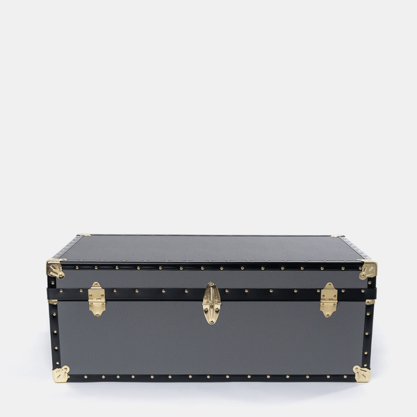 A black leather or faux-leather trunk with gold-tone metal hardware, corner protectors, and rivets, photographed against a plain light gray background.