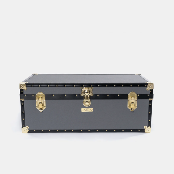 A black vintage-style trunk or storage chest with gold-tone metal hardware, corner protectors, and rivets, photographed against a light gray studio background.
