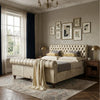 A tufted cream velvet bed with rolled headboard and footboard, styled in a classic bedroom with dark blue curtains, framed botanical wall art, bedside tables with gold lamps, and a patterned rug.