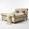 A classic tufted sleigh bed in a warm beige fabric, styled with matching beige bedding, two dark gray pillows, and two gold patterned accent pillows. A beige throw blanket with fringe is draped over the rolled side of the bed, which stands on dark wooden legs against a clean white studio background.