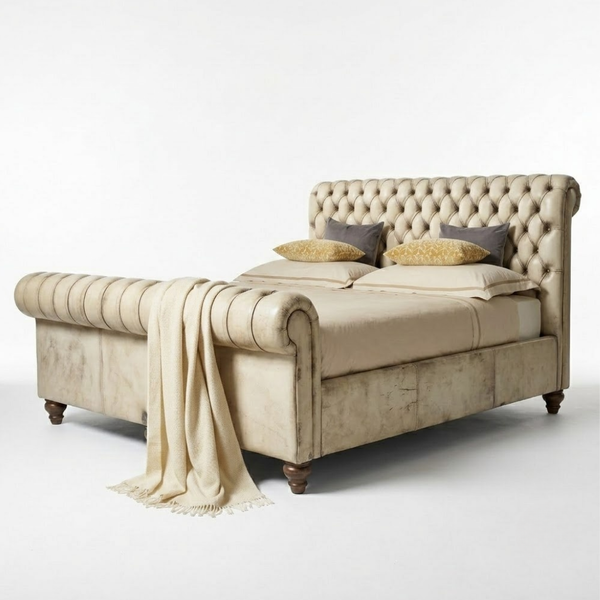 A classic tufted sleigh bed in a warm beige fabric, styled with matching beige bedding, two dark gray pillows, and two gold patterned accent pillows. A beige throw blanket with fringe is draped over the rolled side of the bed, which stands on dark wooden legs against a clean white studio background.