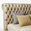 A close-up of a tufted leather bed headboard in a warm taupe-beige color, styled with neutral gray and textured mustard-yellow pillows on a light beige bed against a white background.