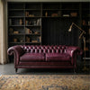 A classic Chesterfield loveseat sofa upholstered in deep burgundy leather, positioned in a dark, elegant room with a large bookshelf and a patterned rug.