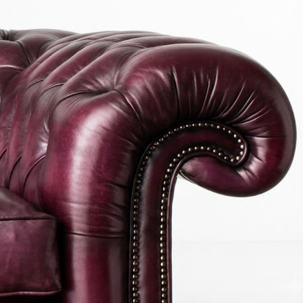 Close-up of a tufted leather armchair in deep burgundy with rolled arms and nailhead trim, photographed against a plain white background.
