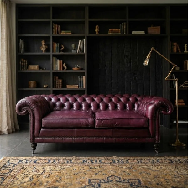 A classic Chesterfield loveseat sofa upholstered in deep burgundy leather, positioned in a dark, elegant room with a large bookshelf and a patterned rug.