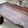 A rustic live-edge wooden table made of dark brown wood with prominent grain patterns, photographed in an outdoor setting against a stone wall.