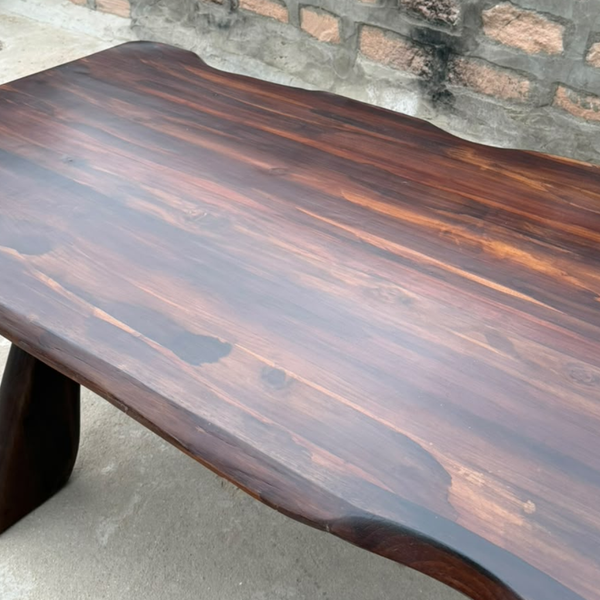 A rustic live-edge wooden table made of dark brown wood with prominent grain patterns, photographed in an outdoor setting against a stone wall.