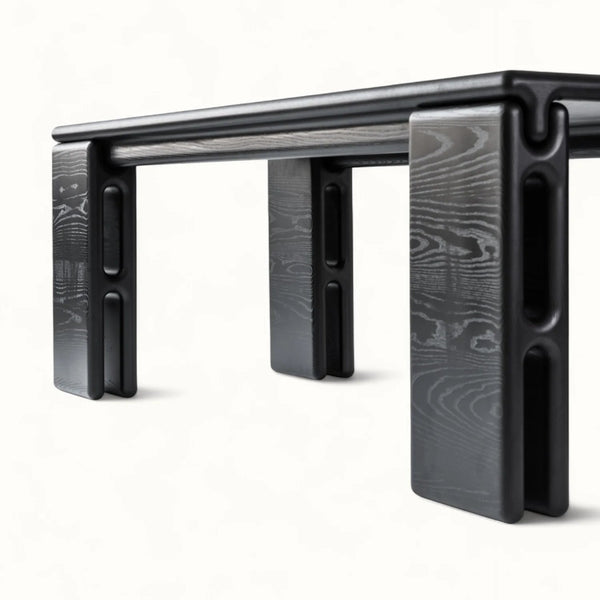 A close-up studio shot of a modern black table with a dark wood grain finish, showing its unique blocky legs and top edge against a clean white background.